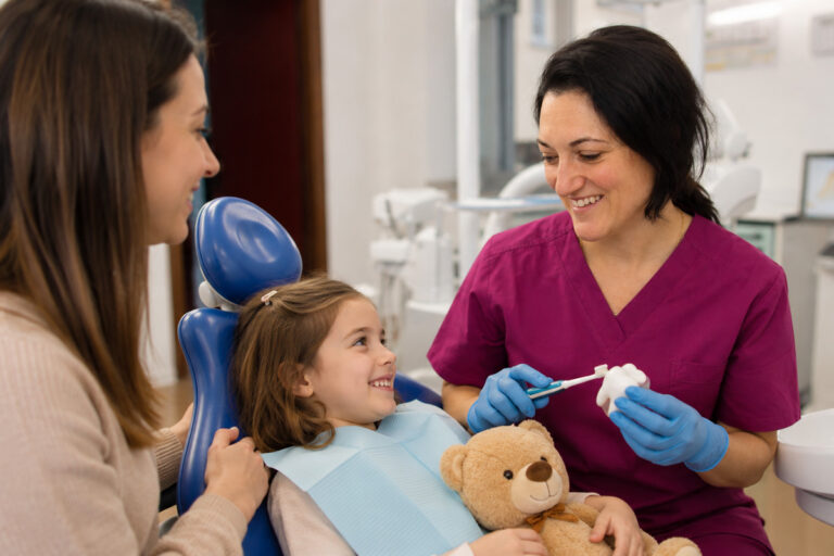 Una bambina in poltrona con il suo orsacchiotto, accanto alla mamma e all'odontoiatra